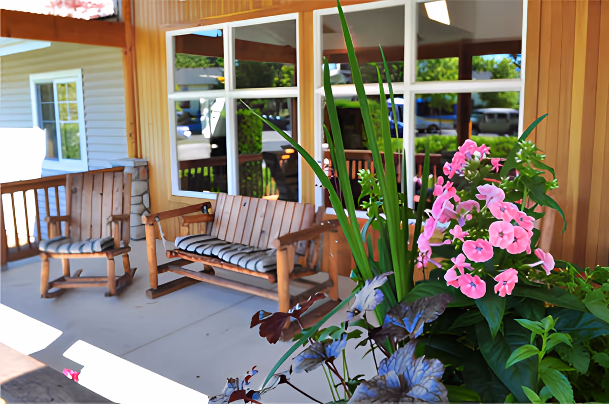 Covered porch with wooden rocking chairs and a bench next to pink flowering plants in front of windows.