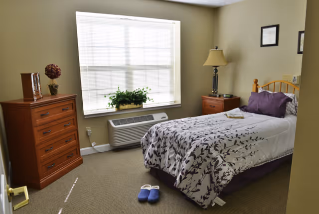 A neatly arranged bedroom with a single bed covered in a white and purple floral bedspread, a wooden nightstand with a lamp, a wooden dresser with decorative items, a window with blinds, and a pair of blue slippers on the carpeted floor.
