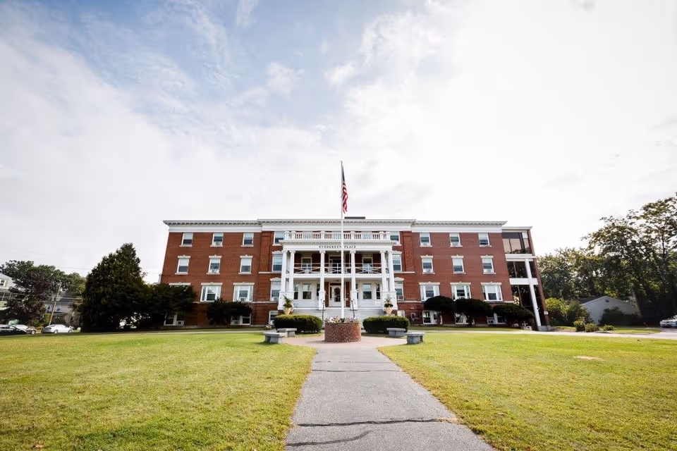 Brick three-story building with a white columned entrance, American flag, and a large front lawn and pathway.