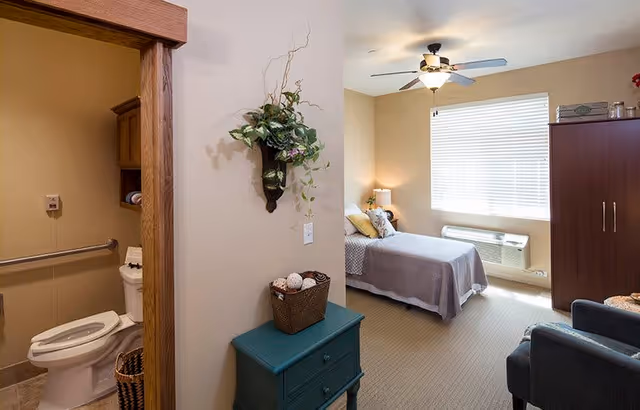 Sunny furnished bedroom with a single bed, wardrobe, armchairs, ceiling fan, and an open adjoining bathroom.