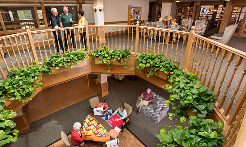 Interior view of a senior living community common area with two levels. On the upper level, three people are standing and talking near a wooden railing decorated with green plants. On the lower level, four seniors are seated around a table working on crafts and reading, with additional seating and bookshelves visible in the background.