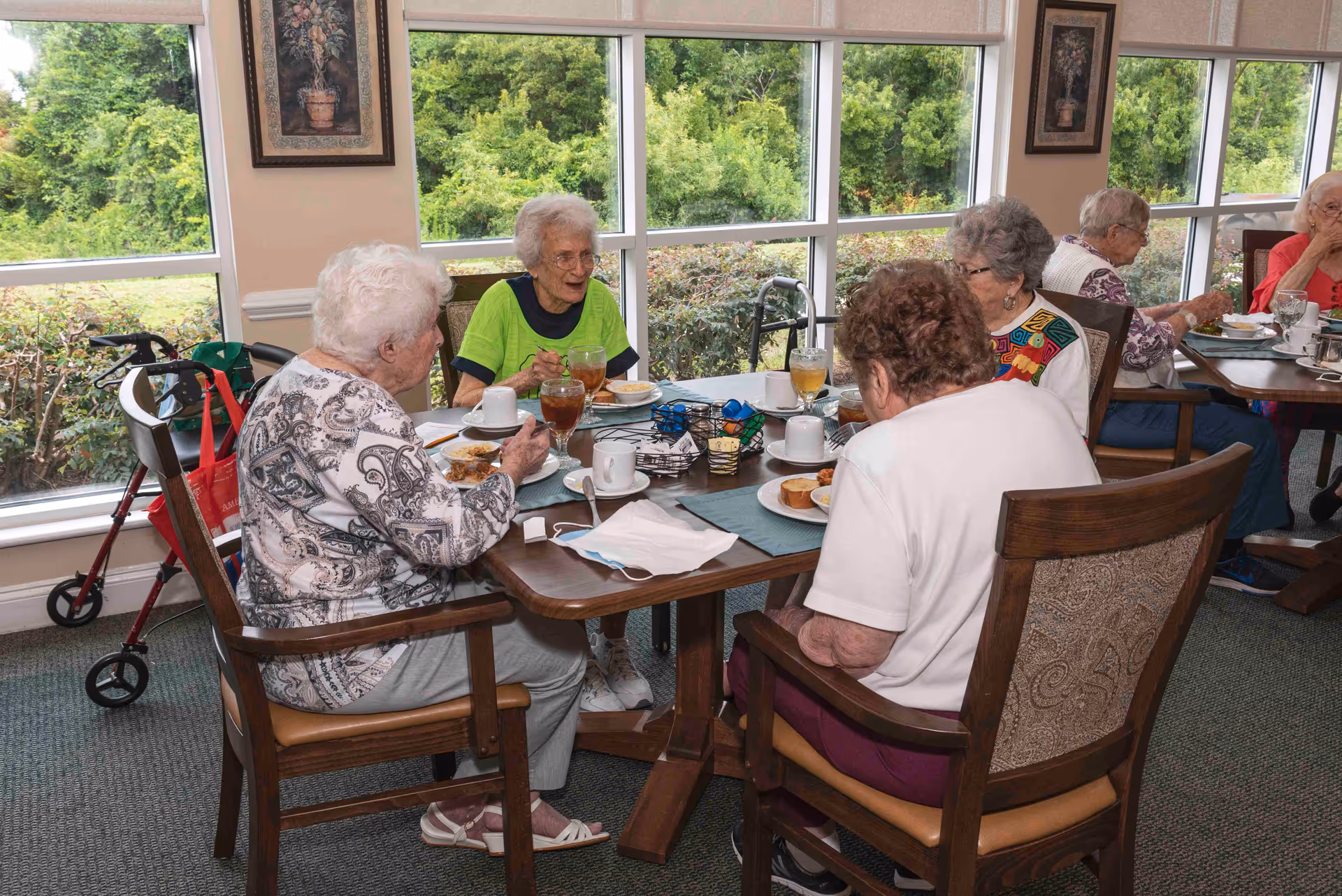 A group of elderly women sitting around a dining table in a well-lit room with large windows showing greenery outside. They are eating and drinking, with plates, cups, and glasses on the table. The room has framed artwork on the walls and comfortable wooden chairs with cushioned seats.