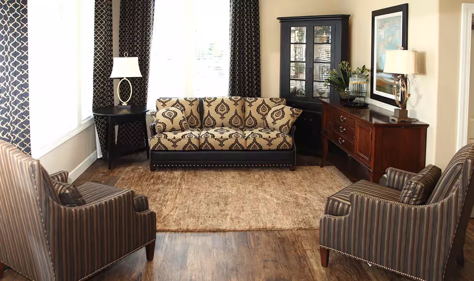 Sunlit sitting area with a patterned sofa and two striped armchairs arranged around a rug, flanked by end tables, lamps, and a wooden sideboard.
