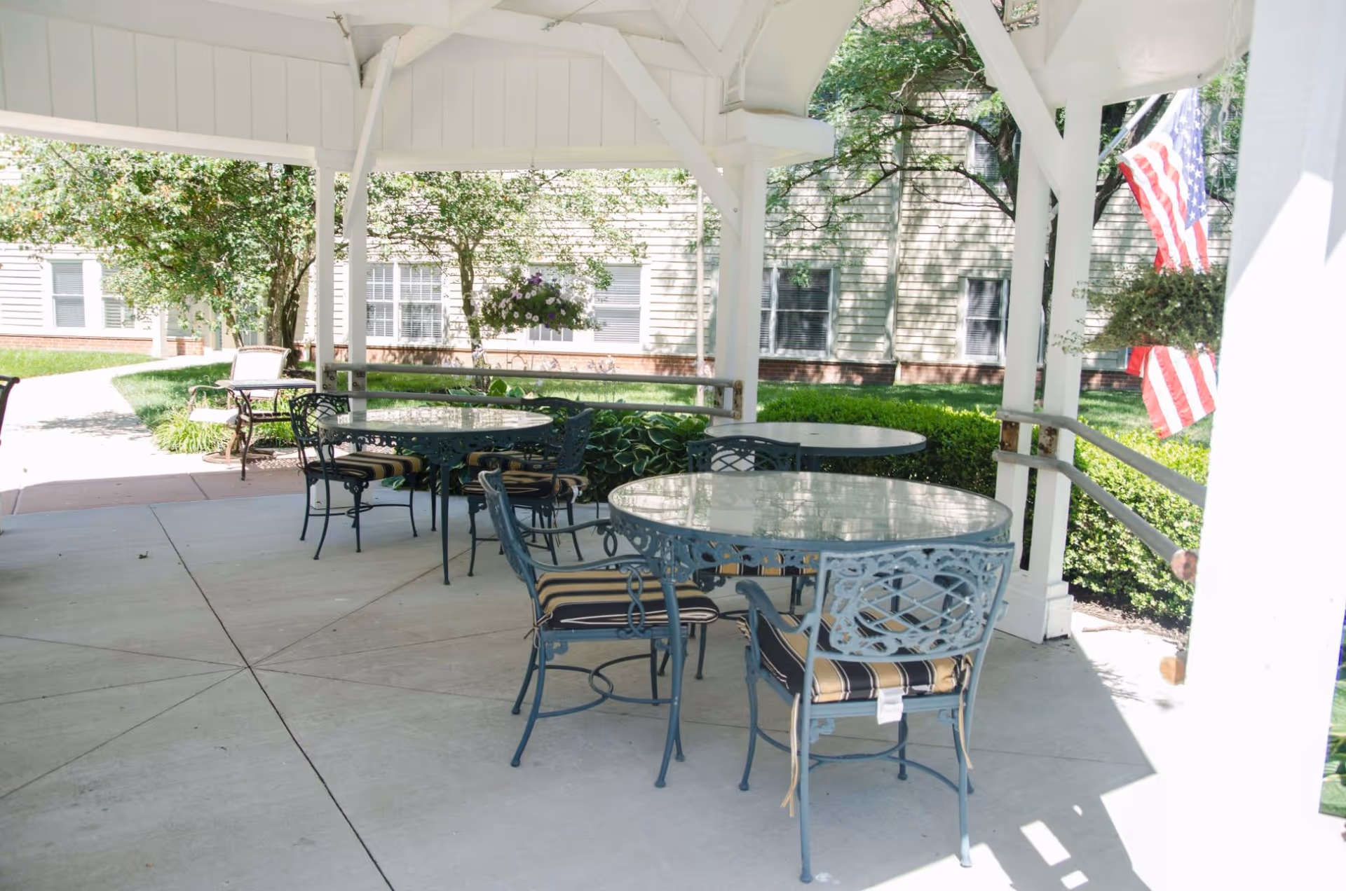 Covered outdoor patio with round glass-top tables and metal chairs under a white gazebo, with landscaping and a building in the background.