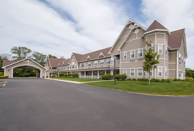 Exterior view of a large, multi-story senior living facility building with beige siding and brown roofing. The building features multiple windows, a covered entrance, and a well-maintained lawn with a few small trees and shrubs. The sky is partly cloudy.