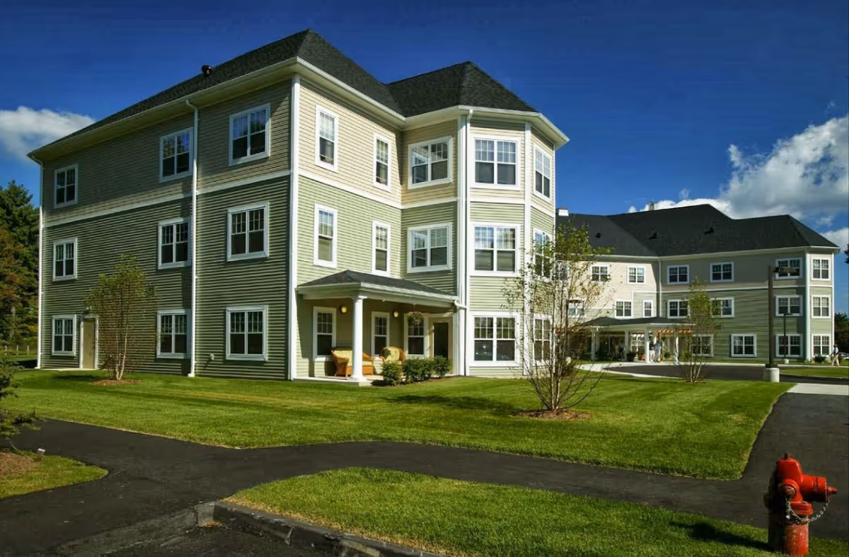 Exterior view of a three-story assisted living facility building with beige and light green siding, multiple windows, a covered entrance with seating, well-maintained green lawns, a paved walkway, and a red fire hydrant in the foreground under a clear blue sky.