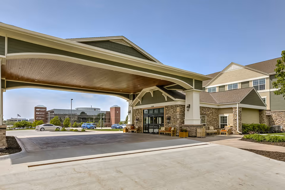 The front entrance and covered drop-off porte-cochere of a multi-story retirement community building with stone accents and outdoor seating.