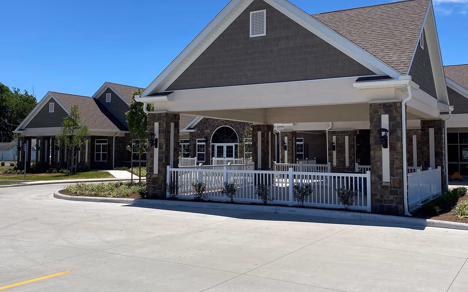 Exterior view of a senior living facility named Rockland Ridge featuring a covered entrance with stone pillars and white railings, surrounded by landscaped greenery and a paved driveway under a clear blue sky.