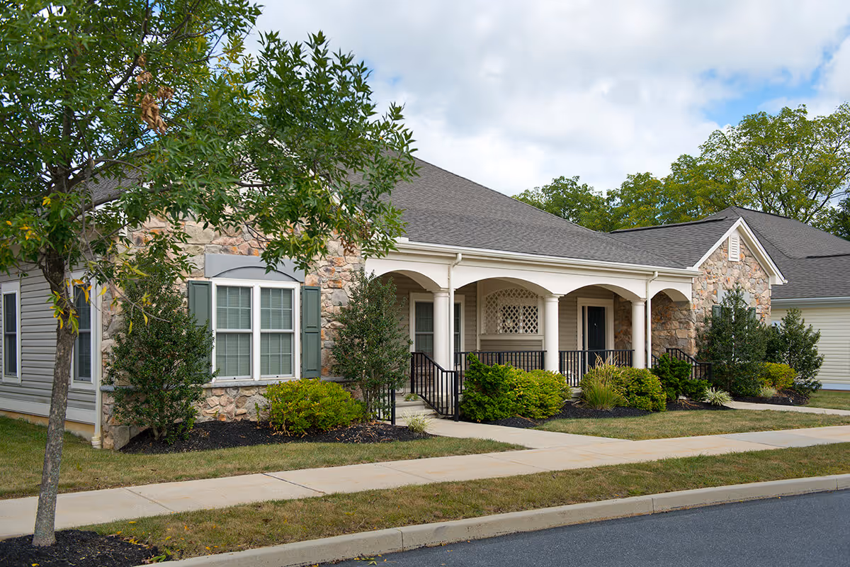 Exterior view of a single-story senior living facility building with stone and siding walls, a covered porch with white columns, green shrubs, and a tree in the front yard under a partly cloudy sky.