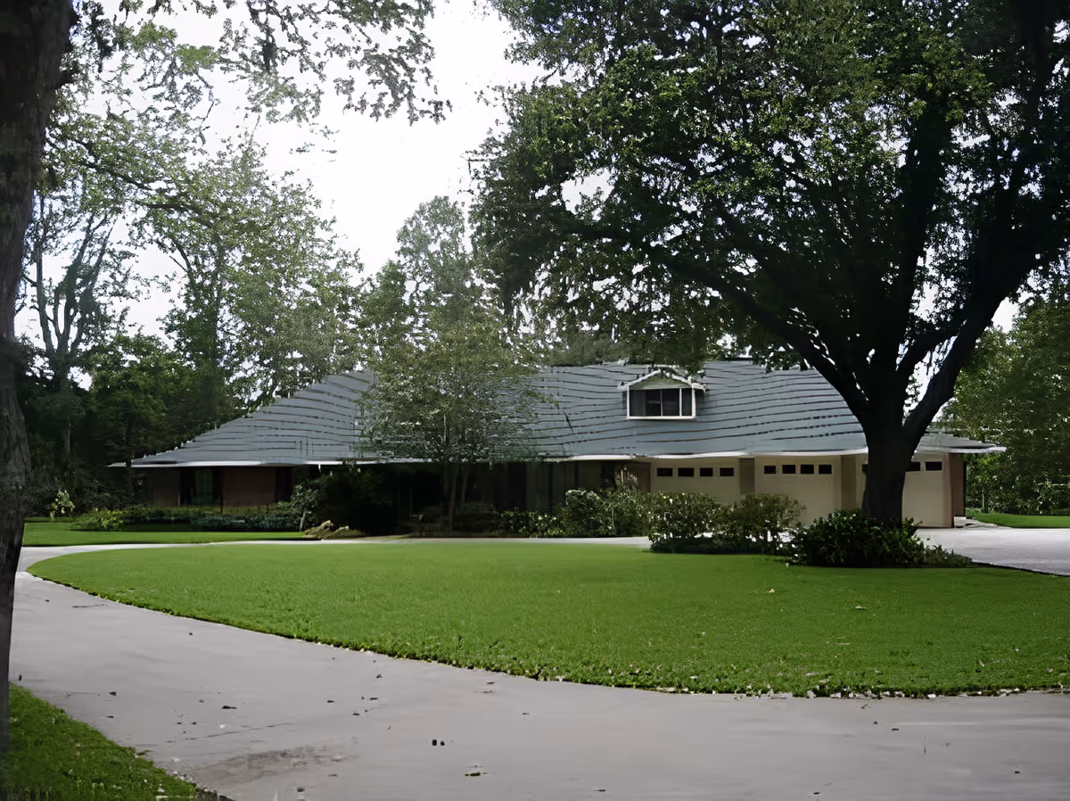A single-story residential building with a gray roof surrounded by green trees and a well-maintained lawn. A curved driveway leads up to the house, which has a garage with multiple doors. Large trees provide shade around the property.