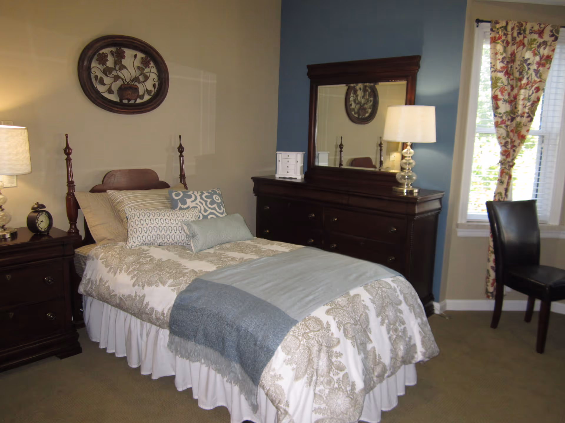 A neatly made single bed with multiple decorative pillows and a patterned bedspread in a bedroom. The room features a wooden nightstand with a lamp and an alarm clock, a large wooden dresser with a mirror and a lamp, a window with floral curtains, and a black chair.