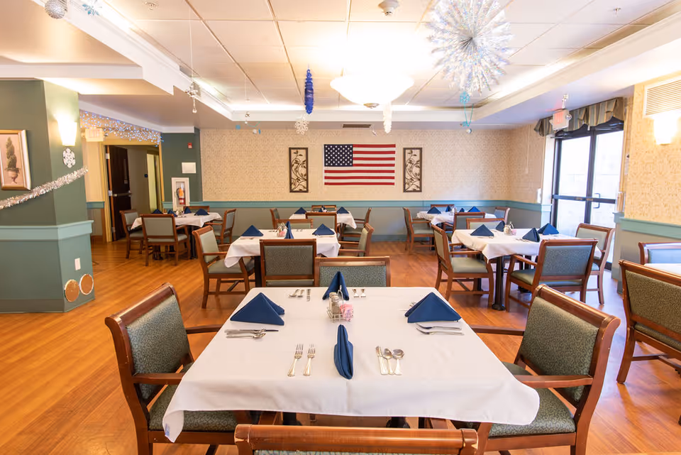 A dining room in a senior living facility with several tables covered in white tablecloths and set with blue folded napkins, silverware, and small flower vases. The room has wooden floors, green and beige walls, and an American flag displayed on the far wall. There are large windows on the right side letting in natural light, and festive decorations hanging from the ceiling.