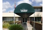 Entrance of Park Ridge Nursing and Rehabilitation facility with a green awning displaying the facility's name. The building is a two-story structure with windows and a pathway leading to double glass doors. There are plants and a small garden area along the walkway, and an American flag is visible on the left side.