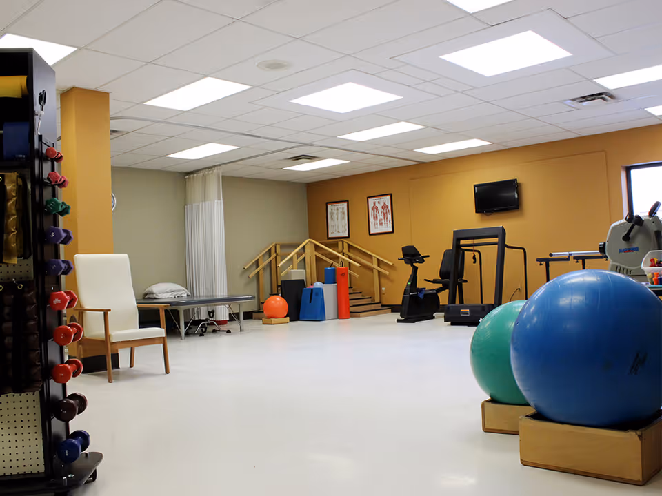 A spacious physical therapy or exercise room with various equipment including colorful dumbbells on a rack, exercise balls, a stationary bike, a treadmill, and a rowing machine. There is a white chair and a medical examination table with a pillow. The room has a beige and light gray color scheme with anatomical charts on the wall and a small set of stairs with handrails.