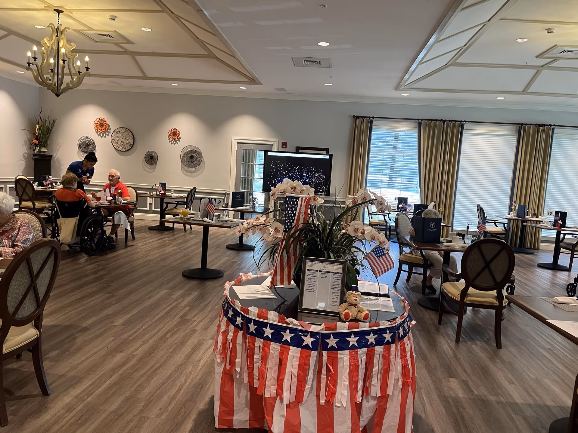 A spacious dining room in a senior living facility decorated with patriotic red, white, and blue streamers and small American flags. Several elderly residents are seated at tables, some in wheelchairs, with a staff member assisting one of them. The room has large windows with beige curtains, wooden flooring, and decorative wall art. A chandelier hangs from the ceiling.