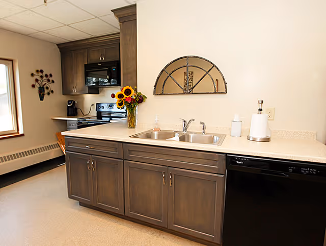 A kitchen area with dark wooden cabinets, a double sink, a dishwasher, and a stove with a microwave above it. There is a vase with sunflowers on the counter and a decorative wall mirror above the sink. A window with a flower decoration is visible on the left side.