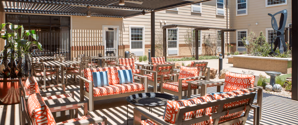 Outdoor patio area at The Legacy at South Plains featuring multiple cushioned chairs and sofas with red and white patterned upholstery, small tables, potted plants, and a modern sculpture in the background. The area is shaded by a pergola casting striped shadows on the furniture and ground.