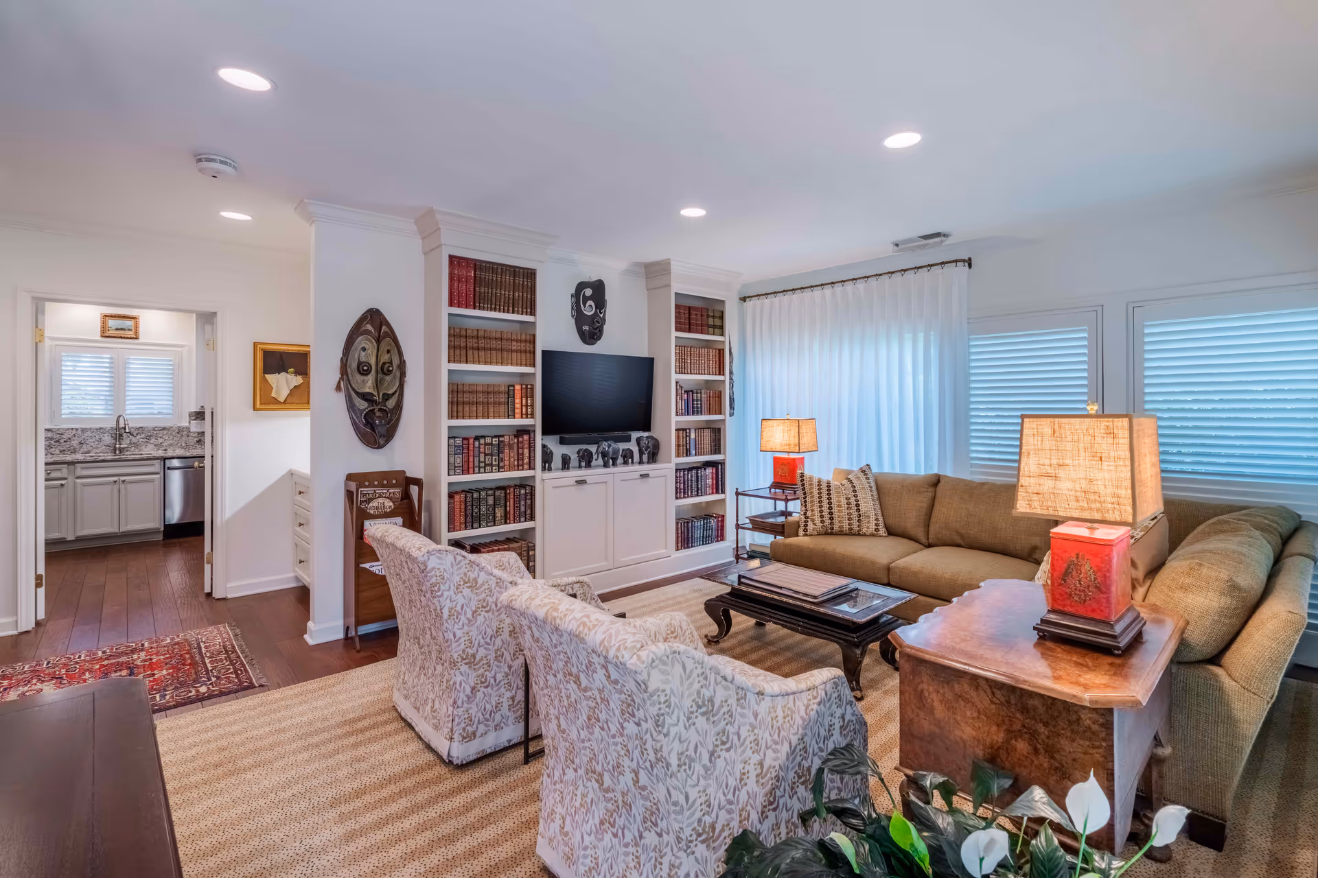 Bright living room with an L-shaped sofa, two patterned armchairs, a coffee table, built-in bookshelves with a TV, and a view into the adjoining kitchen.