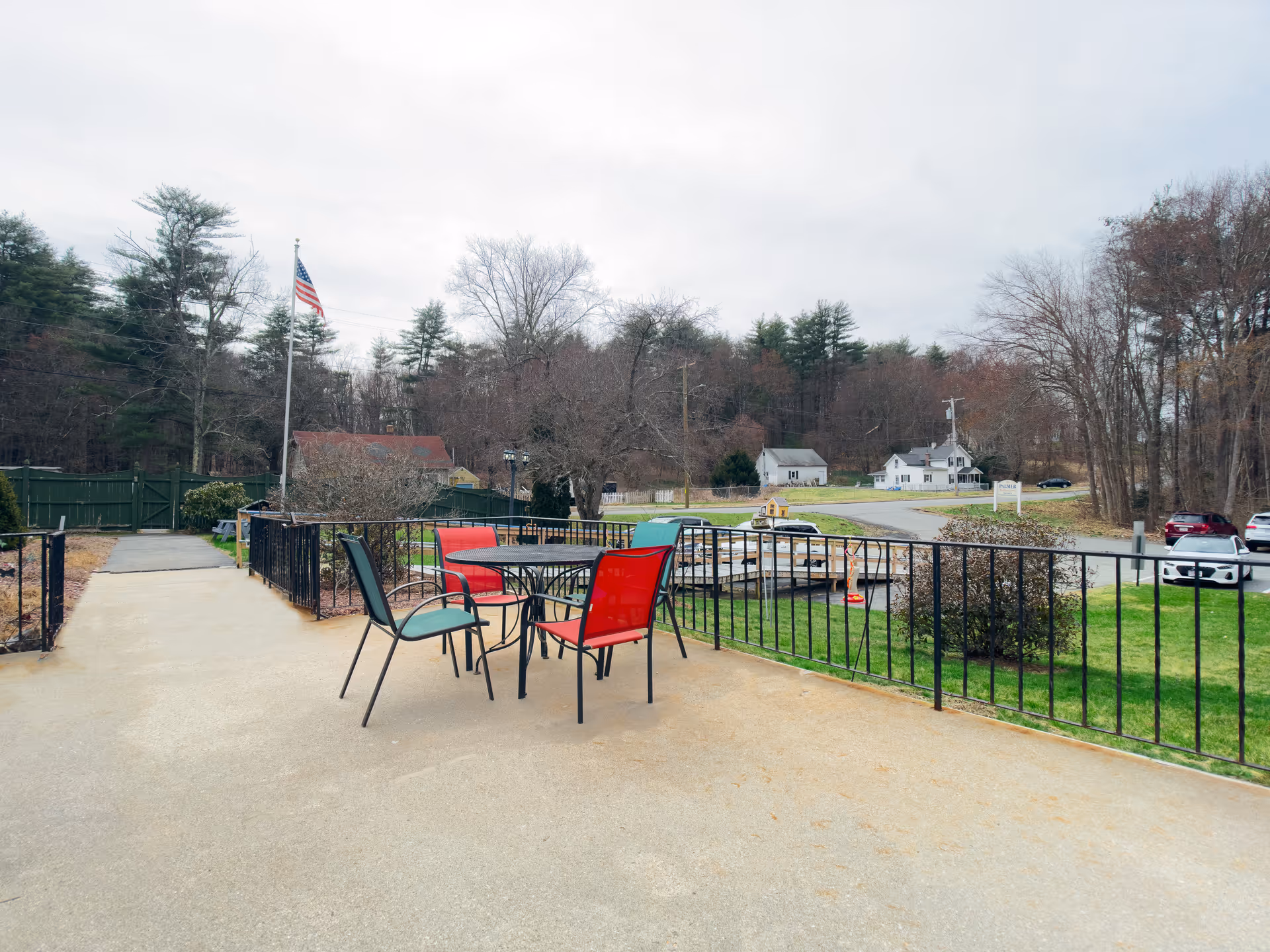 Outdoor patio area with a round table and four chairs, two red and two green, surrounded by a black metal railing. In the background, there is a flagpole with an American flag, trees without leaves, several houses, and parked cars along a road.