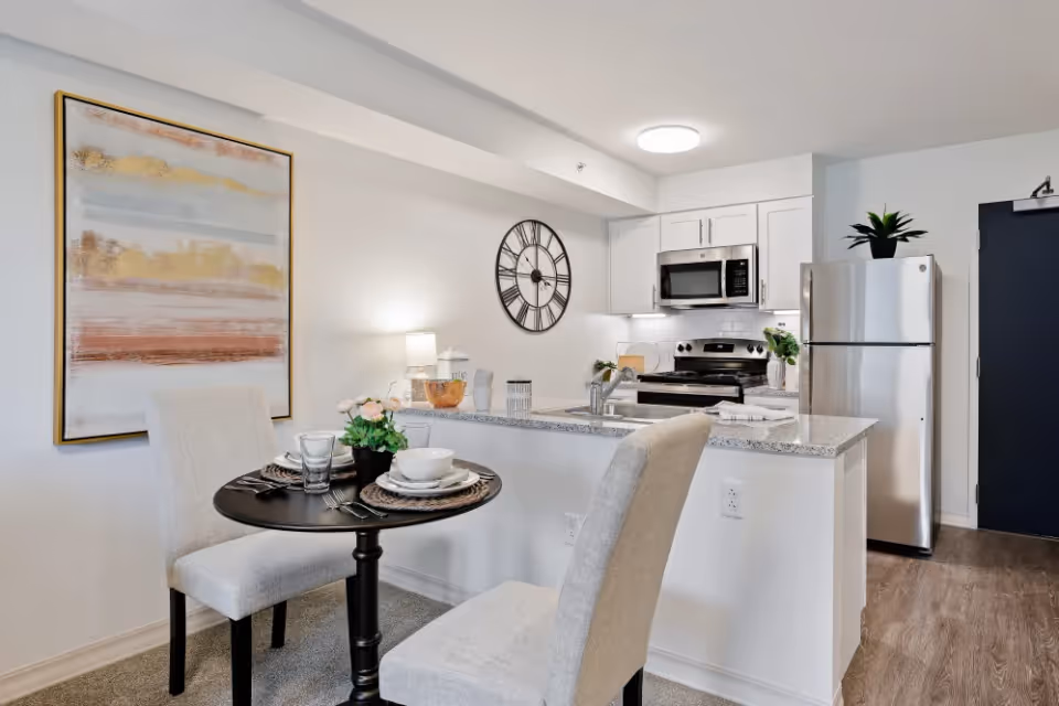 A modern kitchen and dining area in a senior living facility. The kitchen features white cabinets, a stainless steel refrigerator, microwave, and stove. A granite countertop island with a sink separates the kitchen from the dining area, which has a small round black table set for two with beige upholstered chairs. The wall has a large abstract painting and a decorative clock. The flooring is a mix of carpet and wood.