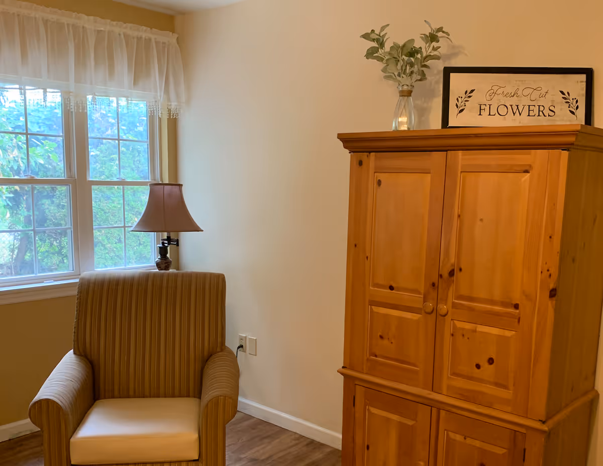Cozy living-room corner with a striped armchair by a window, a table lamp, and a wooden armoire topped with a vase and a 'FLOWERS' sign.