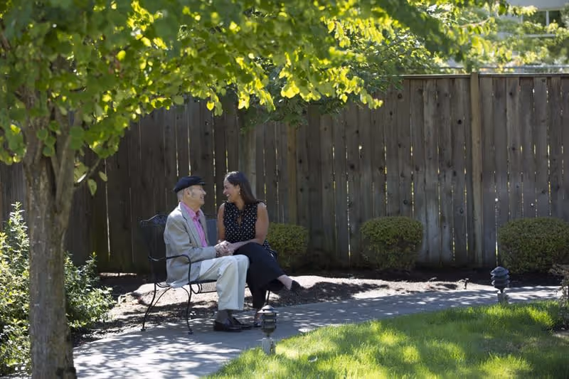 An elderly man and a woman sitting on a metal bench in a garden area, holding hands and smiling at each other. They are surrounded by green grass, bushes, and a wooden fence in the background.