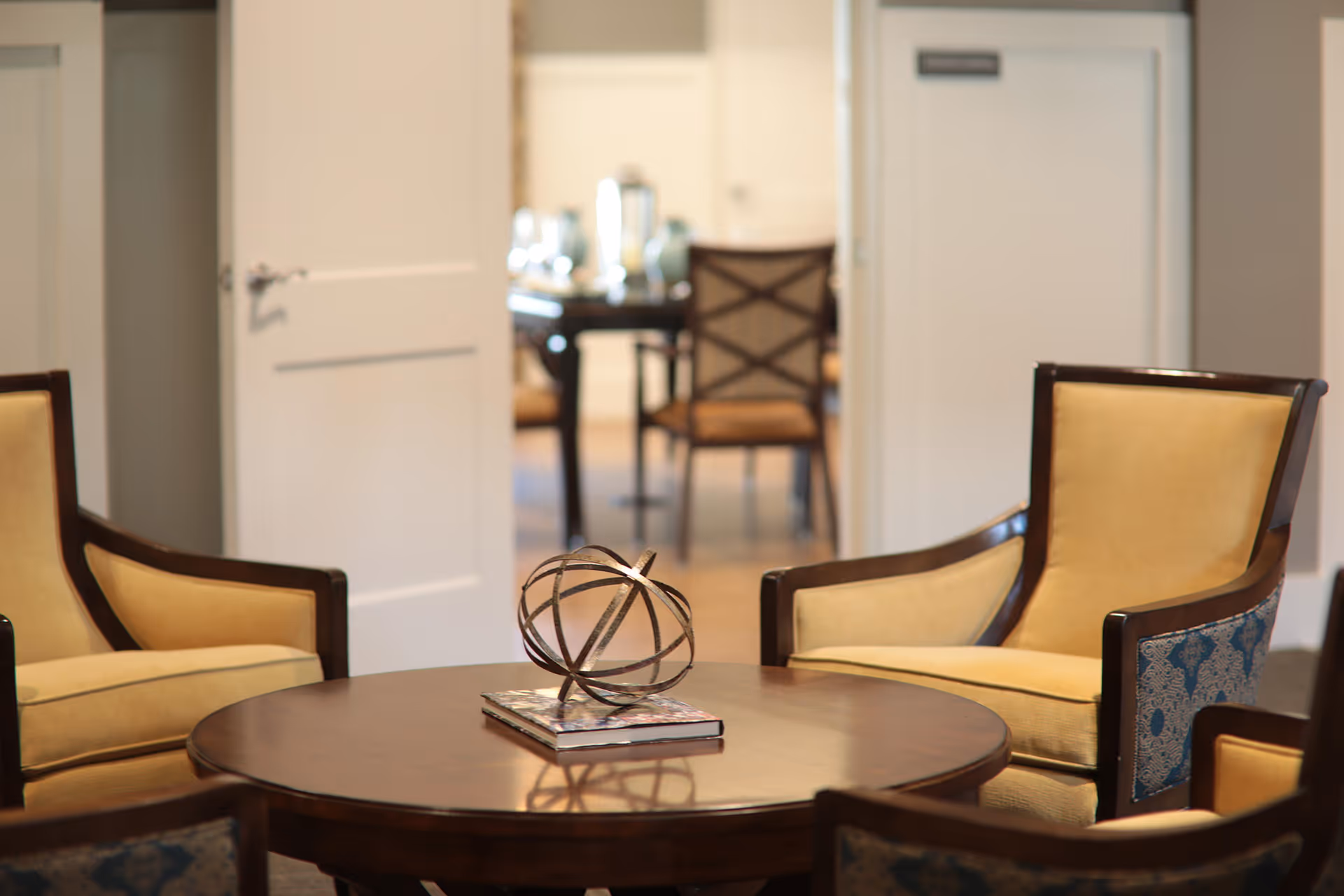 A cozy seating area with four beige armchairs surrounding a round wooden table. On the table, there is a decorative metal orb placed on top of a book. In the background, an open door reveals a dining area with chairs and a table set with glassware.
