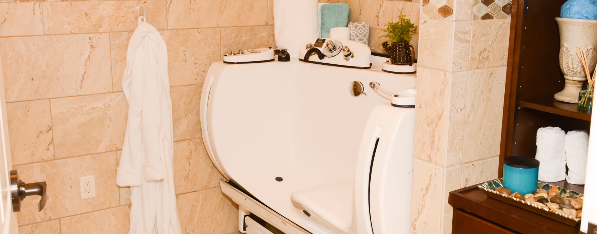 A bathroom with beige tiled walls featuring a white walk-in bathtub equipped with various controls and a built-in seat. A white bathrobe hangs on the wall next to the tub. To the right, there is a wooden shelving unit holding rolled white towels, a decorative vase, a small plant, and a tray with stones and a blue candle.
