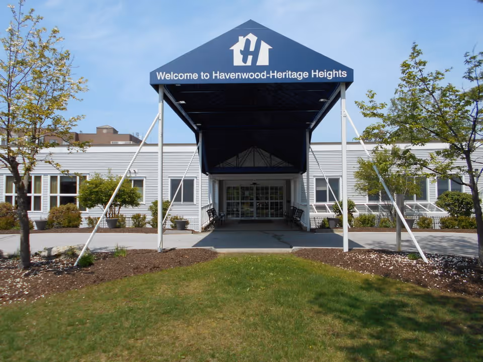 Front exterior view of Havenwood-Heritage Heights facility with a blue canopy entrance that reads 'Welcome to Havenwood-Heritage Heights'. The building is white with several windows and is surrounded by small trees and landscaped greenery.