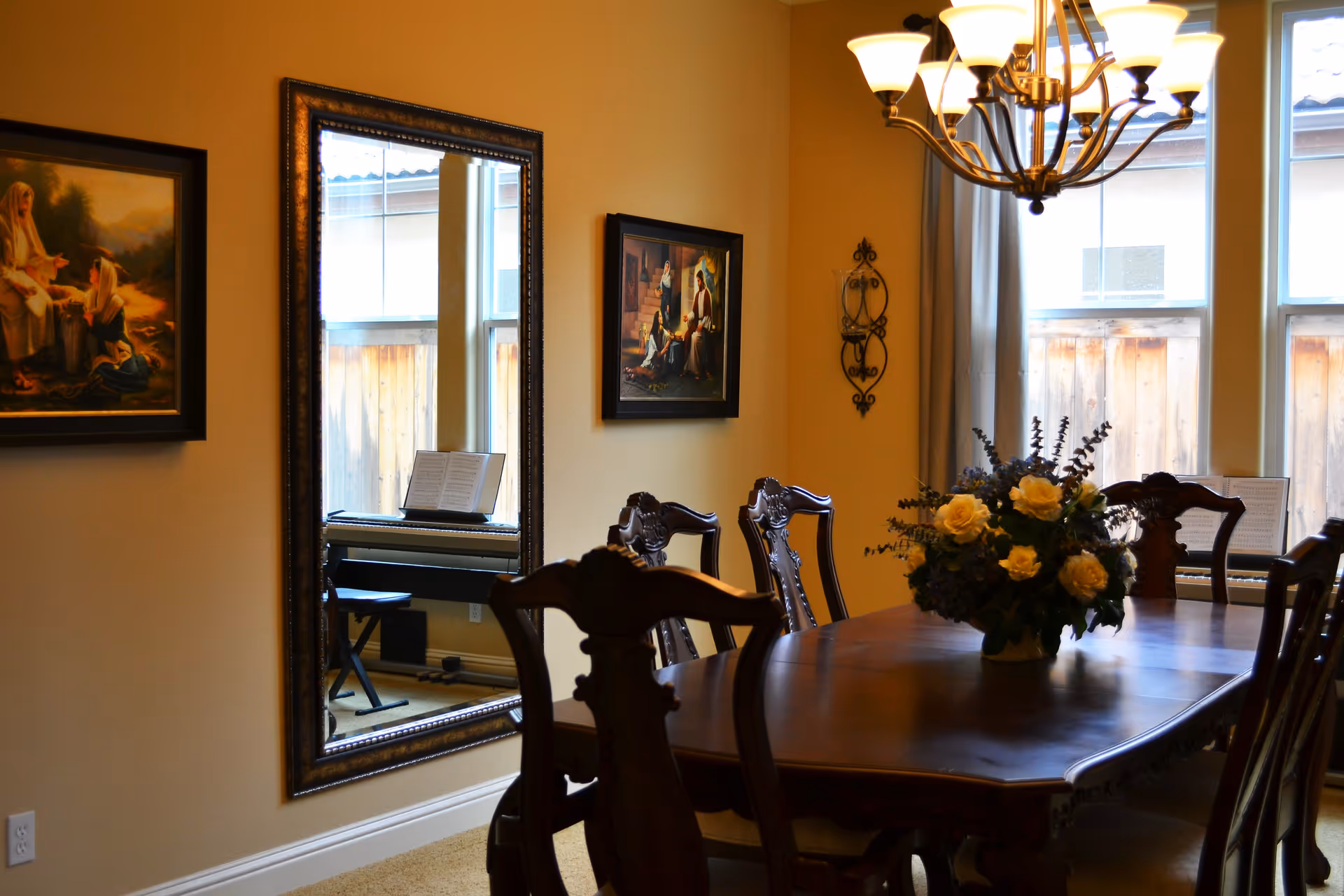 A dining room with a wooden dining table and six chairs. A floral centerpiece with yellow roses is on the table. The room has beige walls with two framed paintings and a large decorative mirror reflecting a piano. There are two windows with curtains letting in natural light, and a chandelier hanging above the table.
