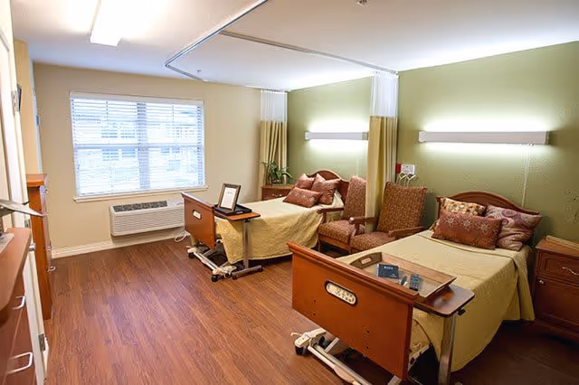A well-lit room with two hospital-style beds, each with a wooden headboard and footboard, beige bedding, and multiple pillows. Between the beds are two upholstered chairs and a small nightstand. The walls are painted green and beige, with a window on the left side letting in natural light. The floor is wood, and there are overhead lights and a ceiling-mounted curtain rail for privacy.