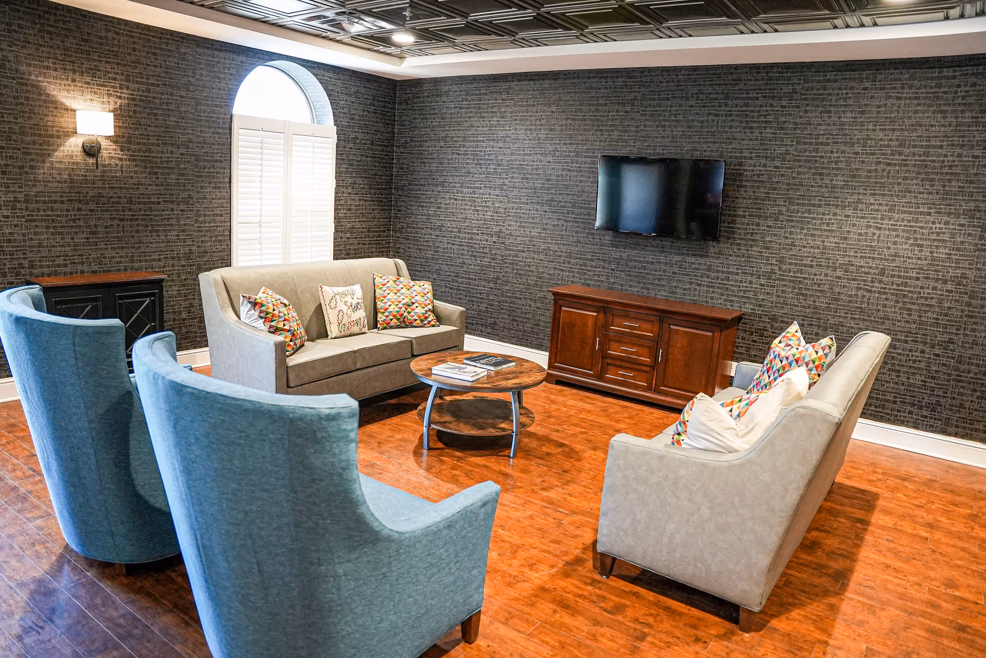 A cozy senior living room with two beige sofas and two blue armchairs arranged around a round wooden coffee table. The room has dark textured wallpaper, a wooden floor, a wall-mounted flat screen TV above a wooden cabinet, and a window with white shutters letting in natural light.