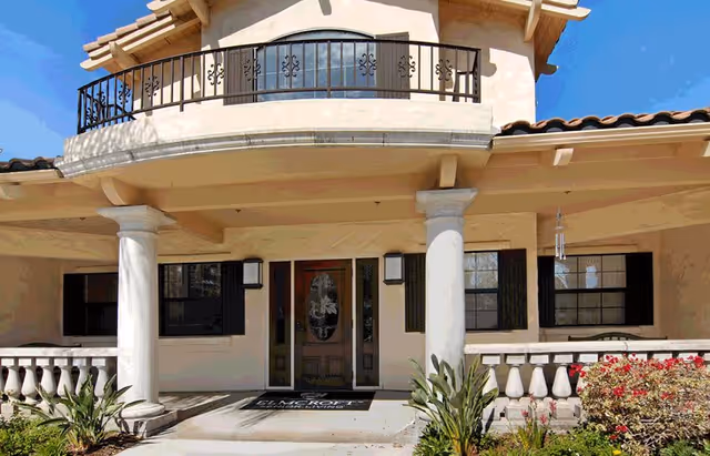 Front exterior view of a two-story building with a covered entrance supported by two large white columns, a balcony with black wrought iron railing above the entrance, and landscaping with plants and flowers in front.