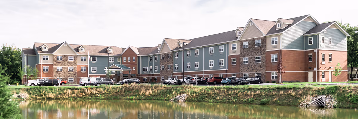 Panoramic exterior view of a three-story senior living building with a pond in the foreground and cars parked along the entrance.