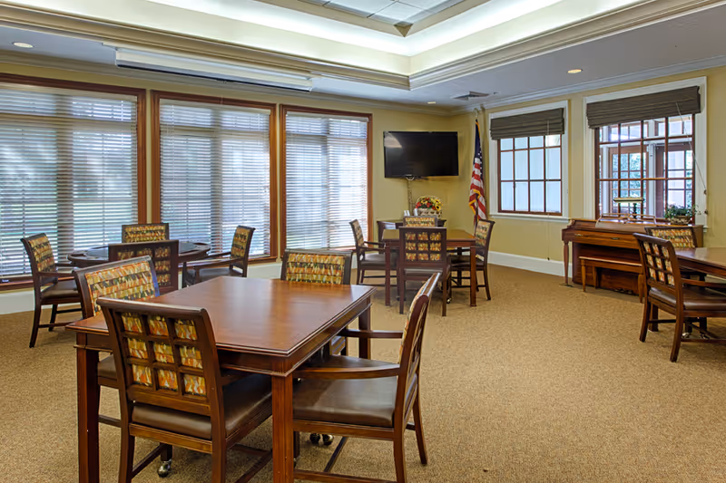 A well-lit room with several wooden tables and chairs arranged for seating. The chairs have patterned cushions. Large windows with blinds cover one wall, and there is a flat-screen TV mounted on the wall next to an American flag. A piano is positioned near another window with blinds.