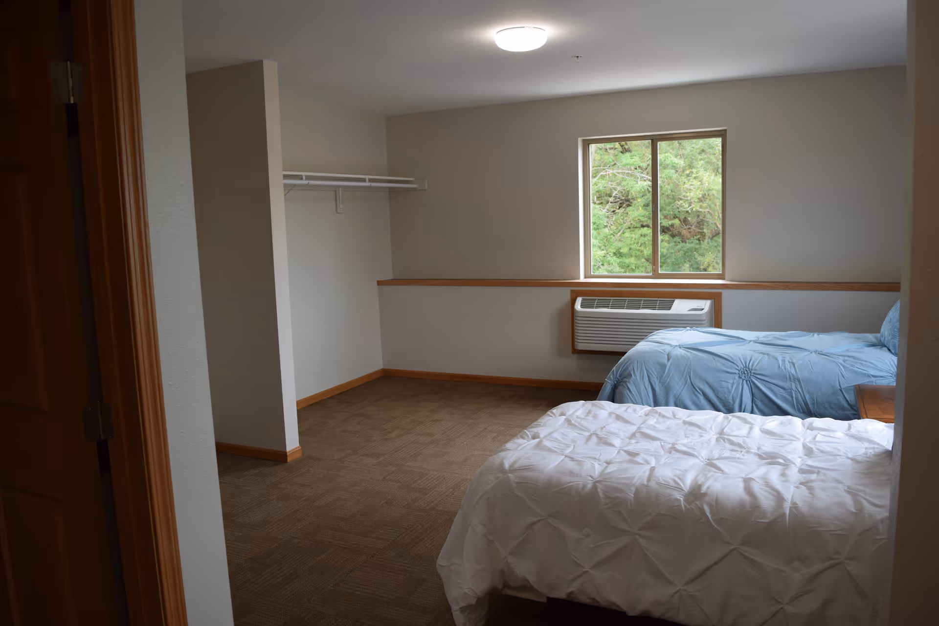 A simple bedroom with two beds, one with a white comforter and the other with a blue comforter. There is a window with a view of greenery outside, an air conditioning unit below the window, and an open closet with a hanging rod on the left wall. The room has beige walls and carpeted flooring.