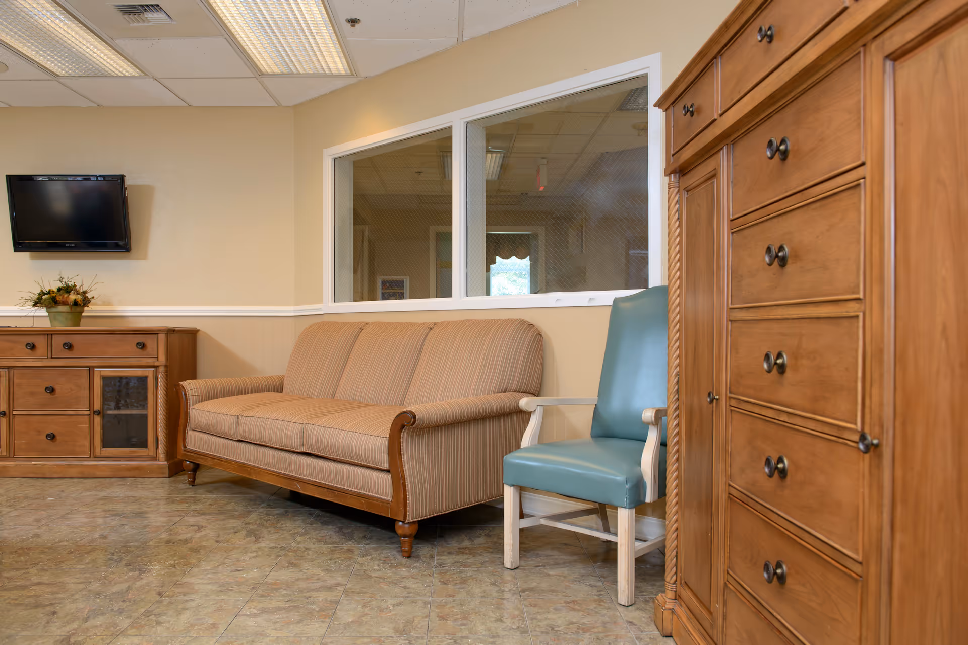 A cozy sitting area in a senior living facility featuring a striped beige sofa, a teal cushioned chair with white wooden arms, a wooden cabinet with drawers and glass doors, a tall wooden chest of drawers, and a wall-mounted flat screen TV. The walls are painted light beige and there is a large window with wire mesh looking into another room.