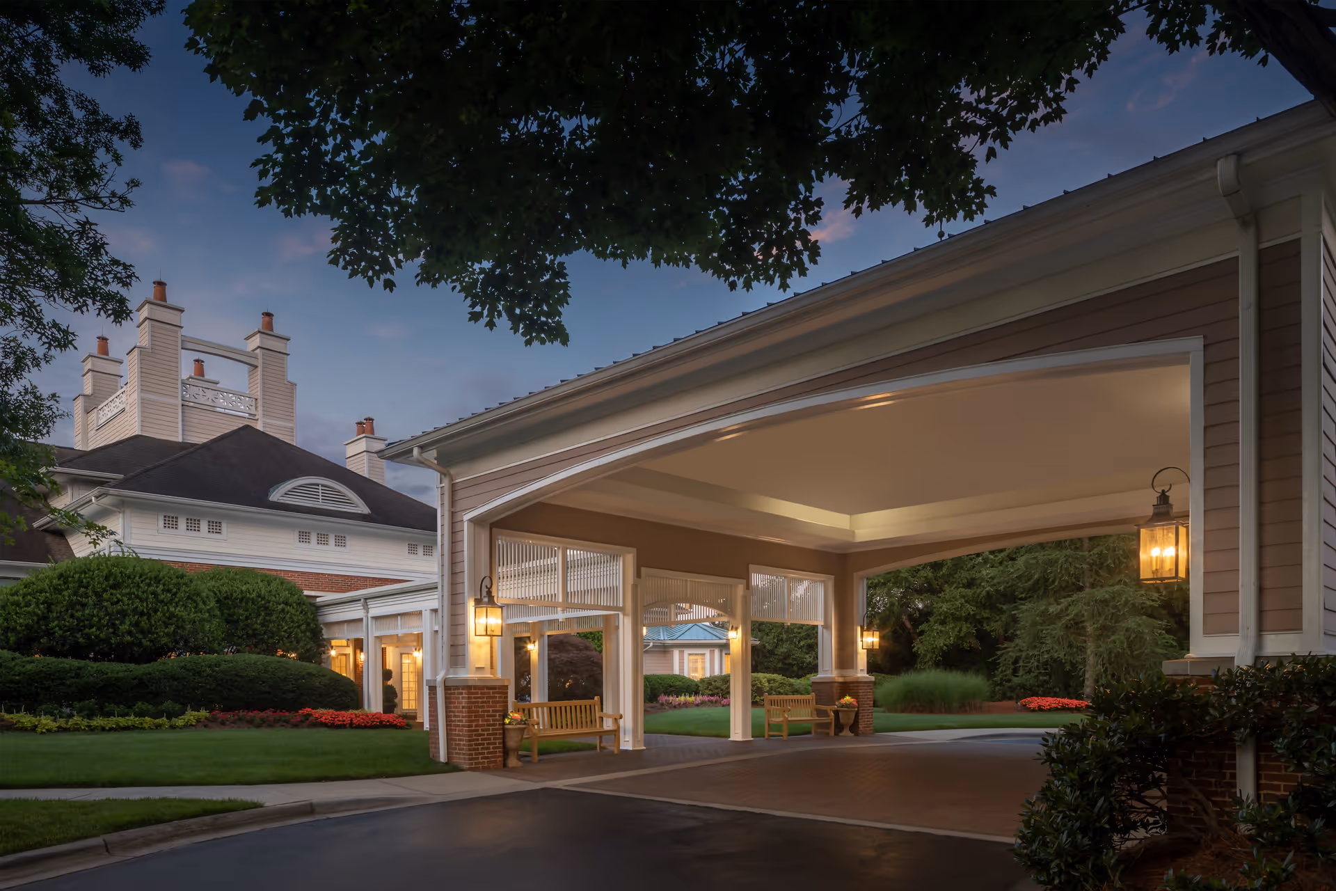 Entrance area of a senior living facility at dusk with a covered drop-off zone featuring hanging lanterns and wooden benches. The building has a classic architectural style with white trim, brick accents, and well-maintained landscaping including bushes and flower beds.
