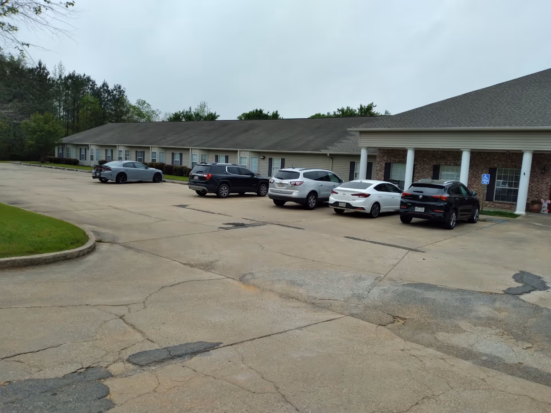 Parking lot in front of a single-story assisted living community building with several cars parked. The building has a brick facade with white columns and multiple windows. Trees and greenery are visible in the background under an overcast sky.