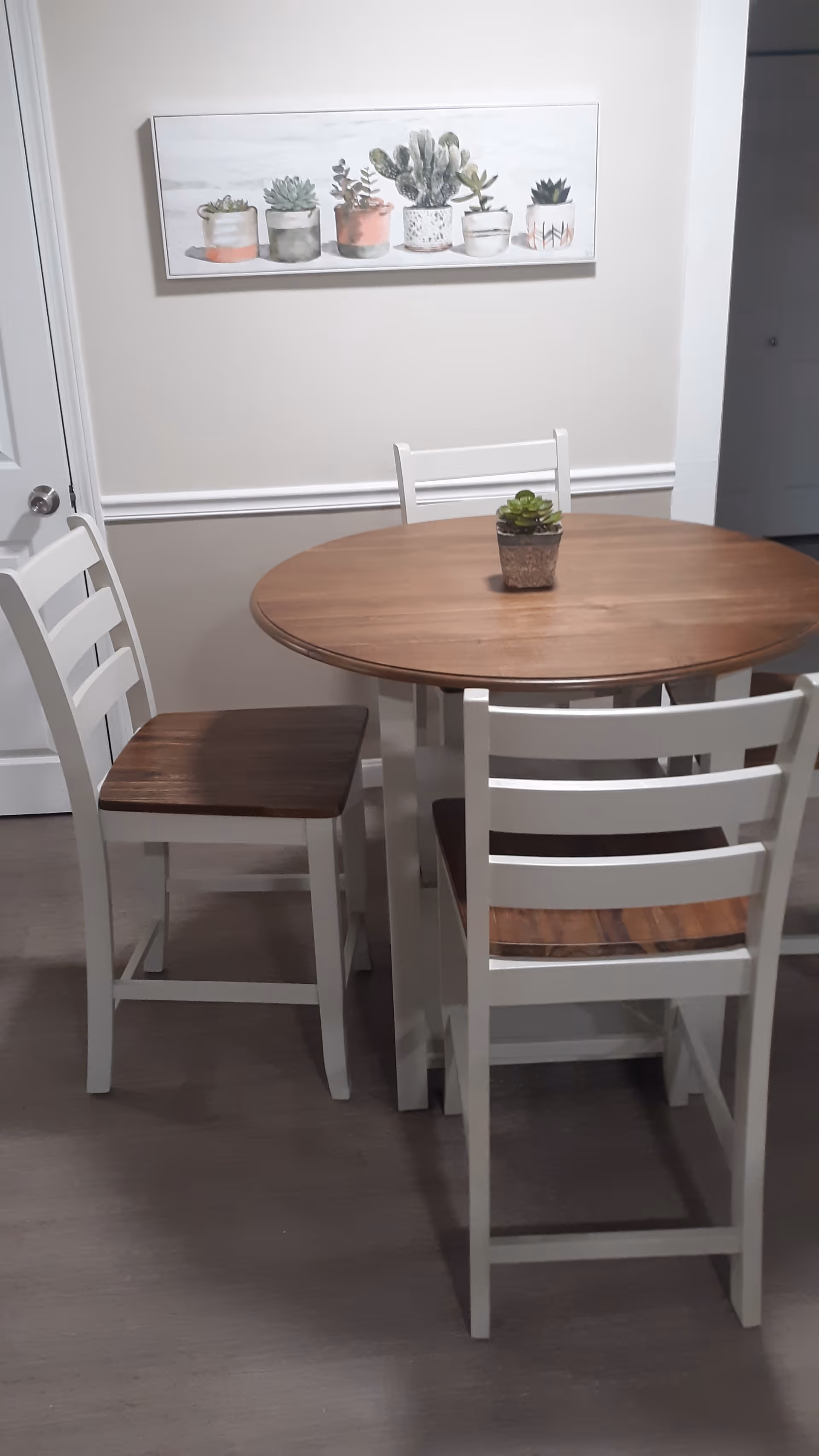 A small round wooden dining table with four white chairs that have wooden seats. A small potted succulent plant is placed in the center of the table. On the wall behind the table, there is a framed picture featuring various potted succulents. The room has light-colored walls and a door to the left.