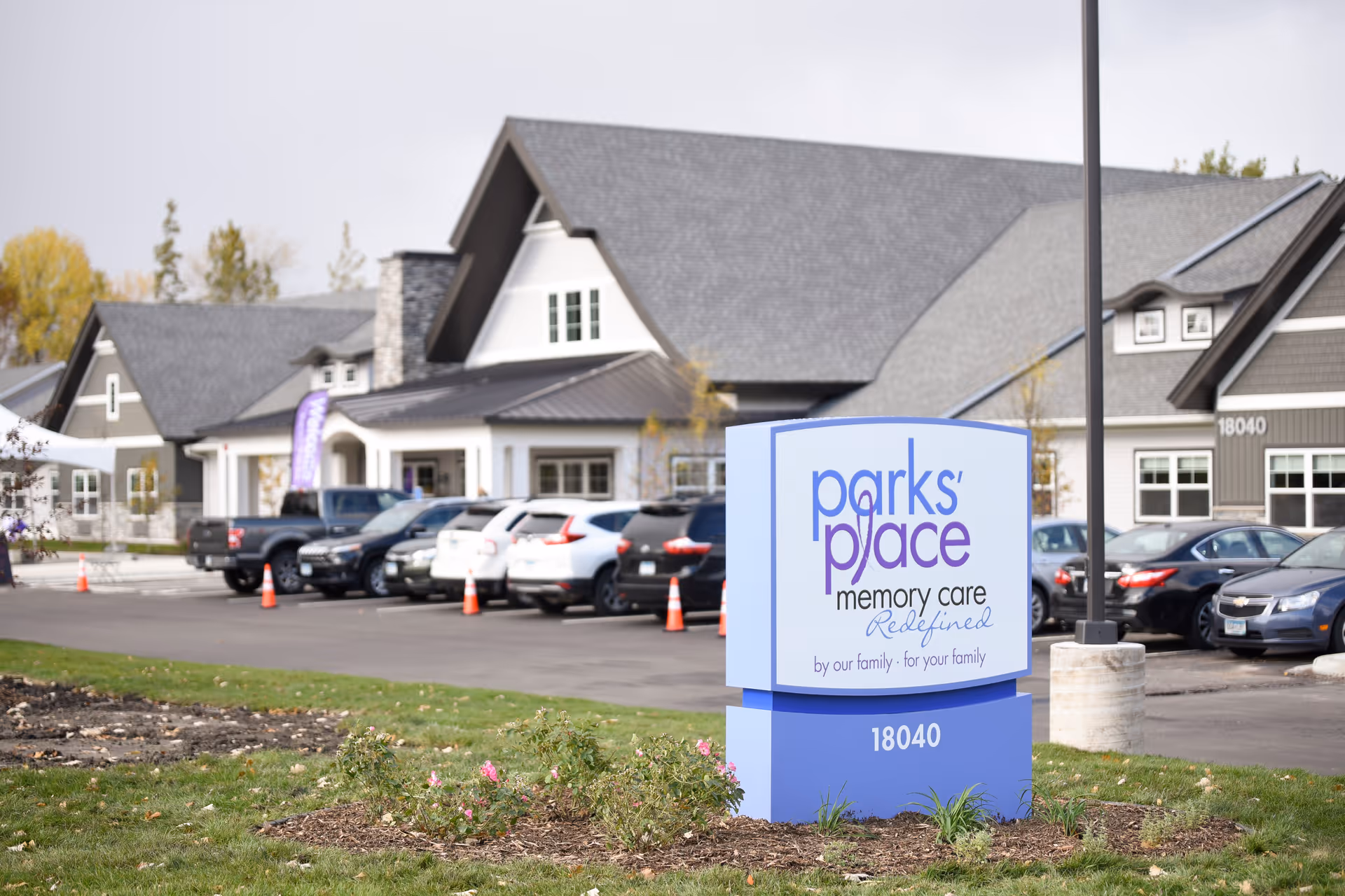 Exterior view of Parks' Place Memory Care facility showing a large sign with the facility name and slogan in front of a parking lot with several cars and a building with gray roofs and white trim in the background.