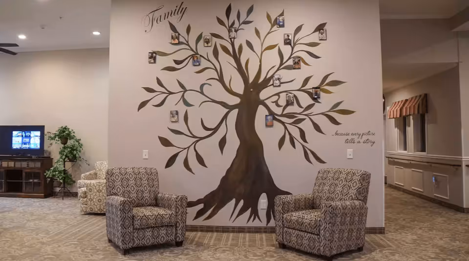 A cozy seating area with two patterned armchairs facing each other in front of a wall painted with a large family tree mural. The mural has small framed photos attached to the branches and the words 'Family' and '...because every picture tells a story' written on the wall. In the background, there is a TV on a wooden stand, a potted plant, and another armchair. The space has soft lighting and a carpeted floor.
