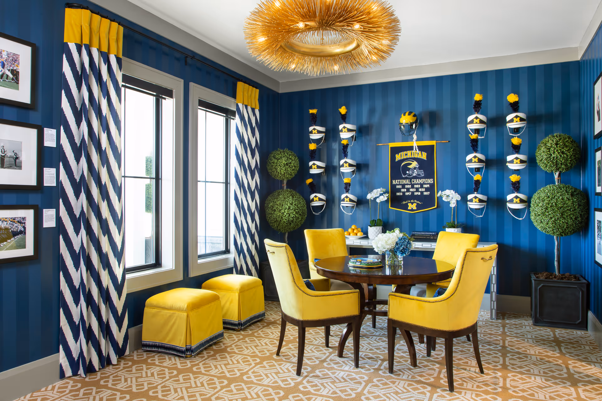 A vibrant room with blue striped walls and yellow accents, featuring a round wooden table surrounded by four yellow upholstered chairs. The room has two windows with blue and white chevron-patterned curtains topped with yellow valances. On the wall, there are multiple Michigan football helmets mounted alongside a Michigan National Champions banner. Two topiary plants in black pots are placed in the corners, and a modern gold chandelier hangs from the ceiling. The floor has a beige and white geometric patterned carpet.