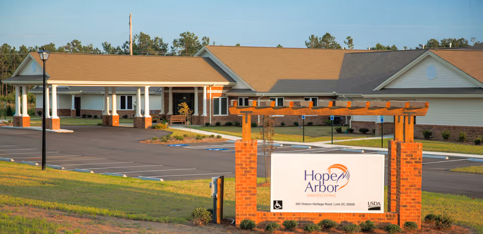 Exterior view of Hope Arbor Of Loris assisted living facility showing a single-story building with a covered entrance, a parking lot with handicap spaces, and a sign in front displaying the facility name and address.
