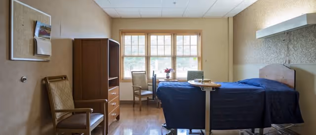 A well-lit bedroom in a senior living facility featuring a hospital-style bed with a dark blue blanket, a wooden headboard, and an adjustable overbed table. The room includes two chairs, a small table with a flower vase near a window with wooden blinds, a wooden dresser with shelves, and a bulletin board on the beige wall.