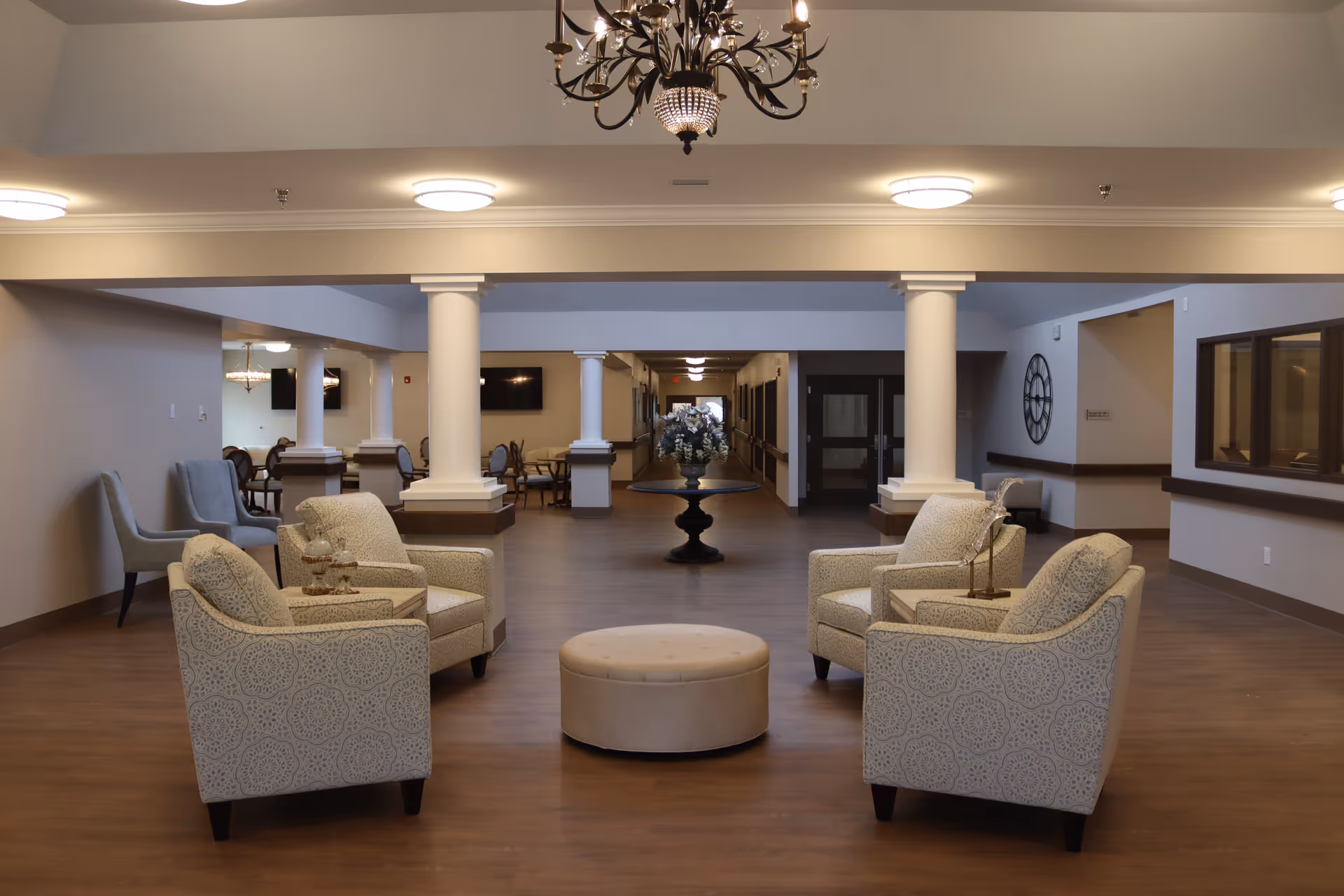 A spacious and well-lit common area in an assisted living facility featuring four patterned armchairs arranged around a round ottoman, with a decorative table holding a floral arrangement in the background. The room has wooden flooring, white columns, and a chandelier hanging from the ceiling.
