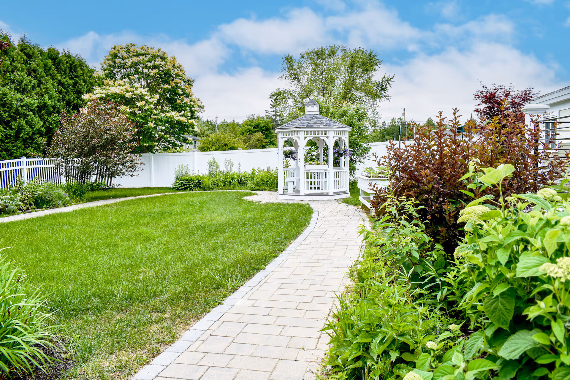 Paved pathway leading through a grassy garden to a white gazebo surrounded by shrubs and trees.