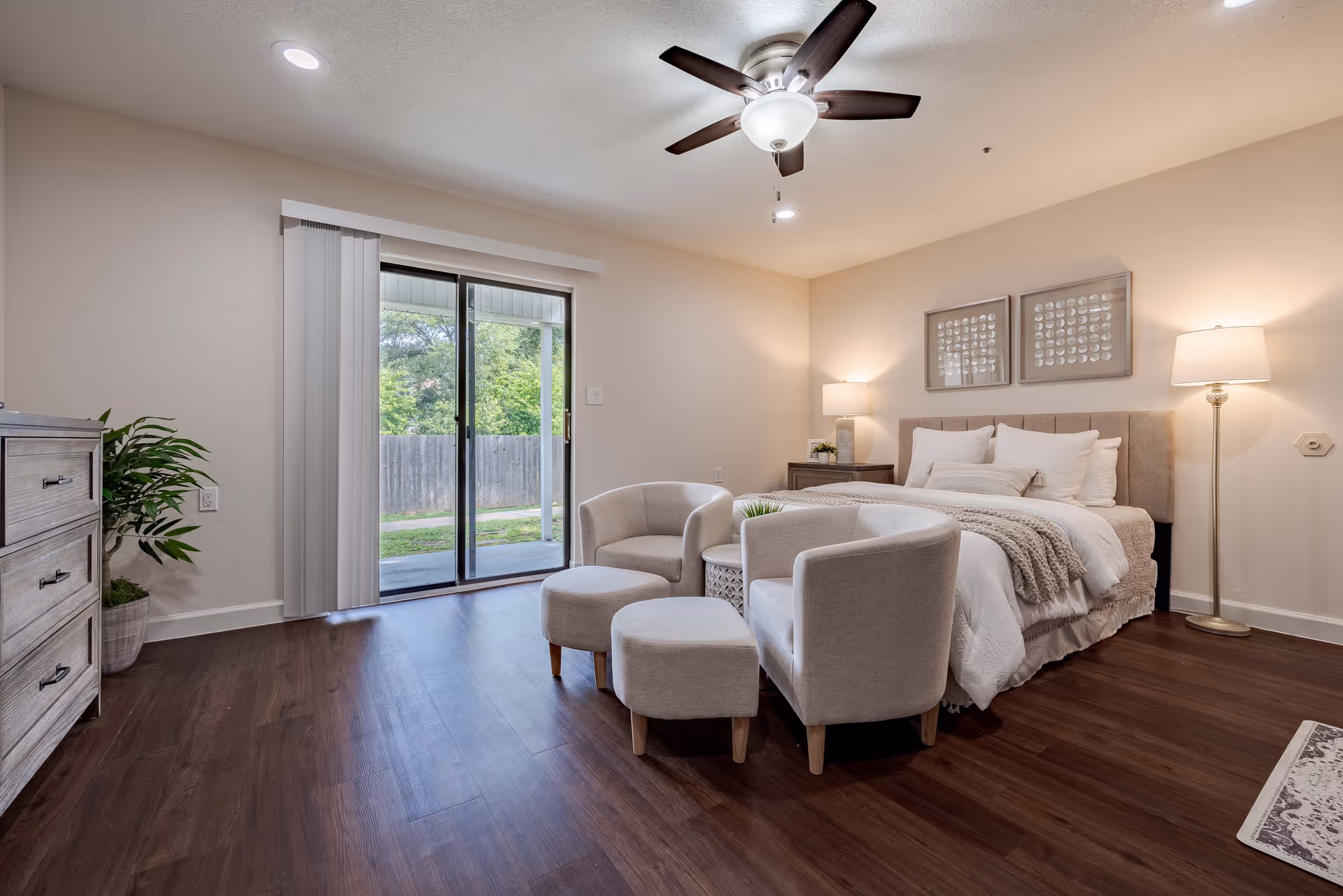 A cozy bedroom with a large bed adorned with white pillows and a beige throw blanket. Two beige armchairs and matching ottomans are placed in front of the bed with a small round table between them. The room has wooden flooring, a ceiling fan with lights, a floor lamp, and a table lamp on a nightstand. Sliding glass doors lead to an outdoor area with greenery visible outside.