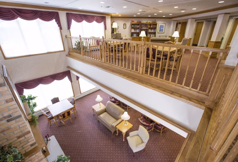 Interior view of a senior living facility common area with two levels. The lower level has a seating area with a sofa, armchair, side tables with lamps, and a table with chairs near large windows with maroon valances. The upper level has more seating with armchairs, a bookshelf, and lamps, surrounded by wooden railings. The space is carpeted with a patterned design and has warm wood accents.