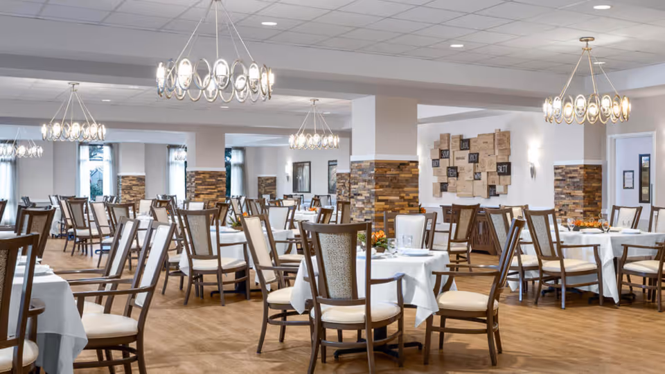 A spacious dining room with multiple round tables covered with white tablecloths, each surrounded by wooden chairs with cushioned seats and backs. The room features modern circular chandeliers hanging from the ceiling, wood-paneled columns, and light-colored walls with decorative artwork. The floor is wooden, and the overall ambiance is bright and inviting.